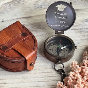 May include: A vintage-style compass with a leather case. The compass is open, revealing the text "BBA 2022 So proud of your work Amazing job love." The compass has a black chain and a needle pointing north. The leather case is brown.