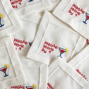 Puede incluir: Una pila de servilletas de c&oacute;ctel de lino blanco con texto bordado en rojo que dice "please leave by 9" y una copa de c&oacute;ctel bordada en rojo y azul con una rodaja de lim&oacute;n amarilla.