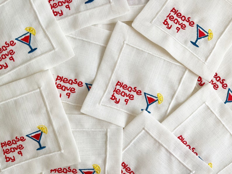 Puede incluir: Una pila de servilletas de c&oacute;ctel de lino blanco con texto bordado en rojo que dice "please leave by 9" y una copa de c&oacute;ctel bordada en rojo y azul con una rodaja de lim&oacute;n amarilla.
