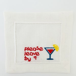 Puede incluir: Servilleta de c&oacute;ctel de lino blanco con bordado rojo que dice "please leave by 9" y una copa de martini roja y azul con una rodaja de lim&oacute;n amarilla.