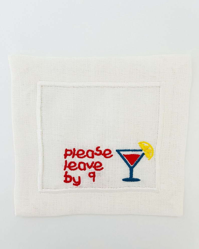 Puede incluir: Servilleta de c&oacute;ctel de lino blanco con bordado rojo que dice "please leave by 9" y una copa de martini roja y azul con una rodaja de lim&oacute;n amarilla.
