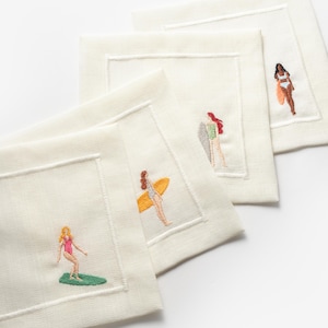May include: Four white linen napkins with embroidered images of women surfing. Each napkin features a different woman in a swimsuit, riding a surfboard with a colourful design.