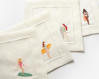 Puede incluir: Cuatro servilletas de lino blanco con imágenes bordadas de mujeres surfeando. Cada servilleta presenta a una mujer diferente en traje de baño, montando una tabla de surf con un diseño colorido.