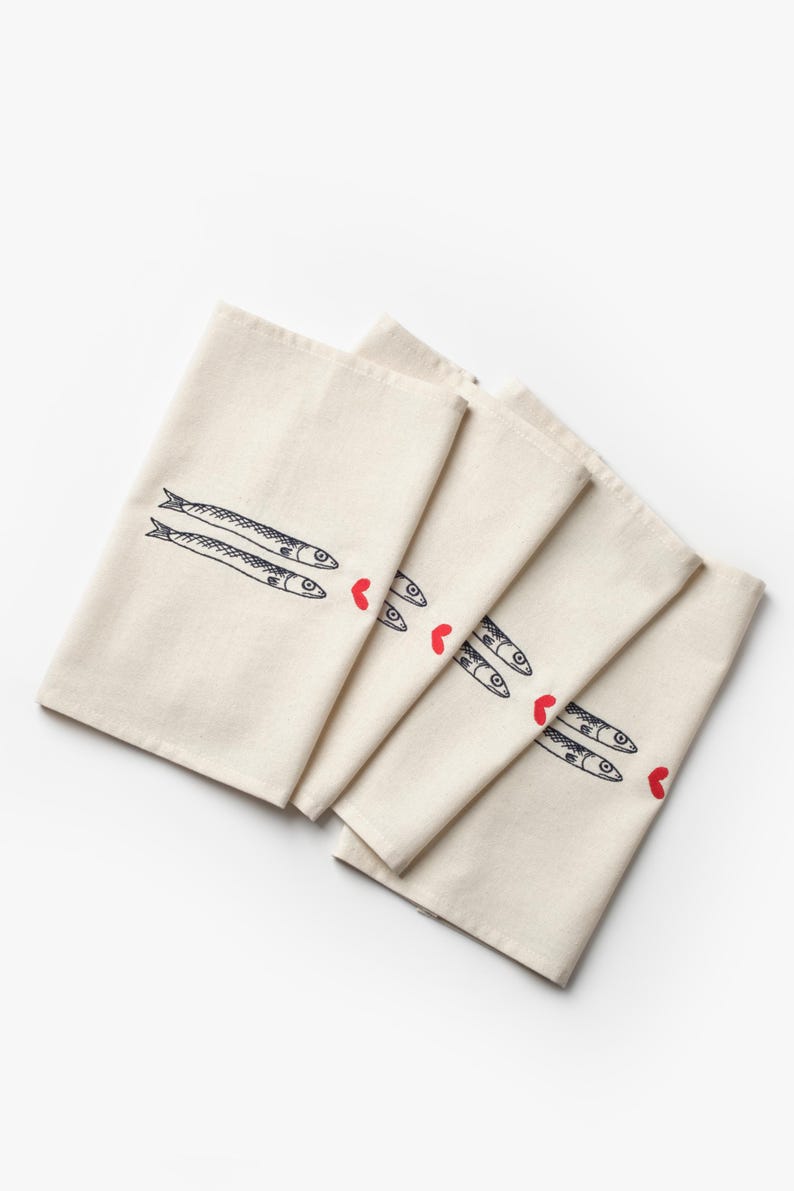 Puede incluir: Pa&ntilde;os de cocina de color crema con dise&ntilde;o de pescado. Cada pa&ntilde;o presenta un par de peces estilizados en tinta negra, con un peque&ntilde;o acento de coraz&oacute;n rojo. Los pa&ntilde;os est&aacute;n doblados y dispuestos en forma de abanico.