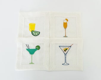 Puede incluir: Conjunto de cuatro servilletas de cóctel de lino blanco con imágenes bordadas de diferentes bebidas: un mimosa, una margarita, un martini y un vaso de zumo de naranja.