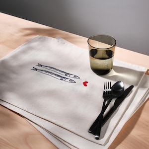 May include: A cream linen placemat featuring a drawing of two fish and a red heart. Black cutlery and a dark glass of water are on the placemat. The scene is set on a light wood table.