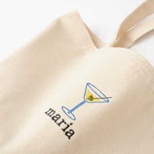 May include: A white canvas tote bag with a blue and yellow martini glass and olive embroidered on the front. The word "maria" is embroidered below the glass.