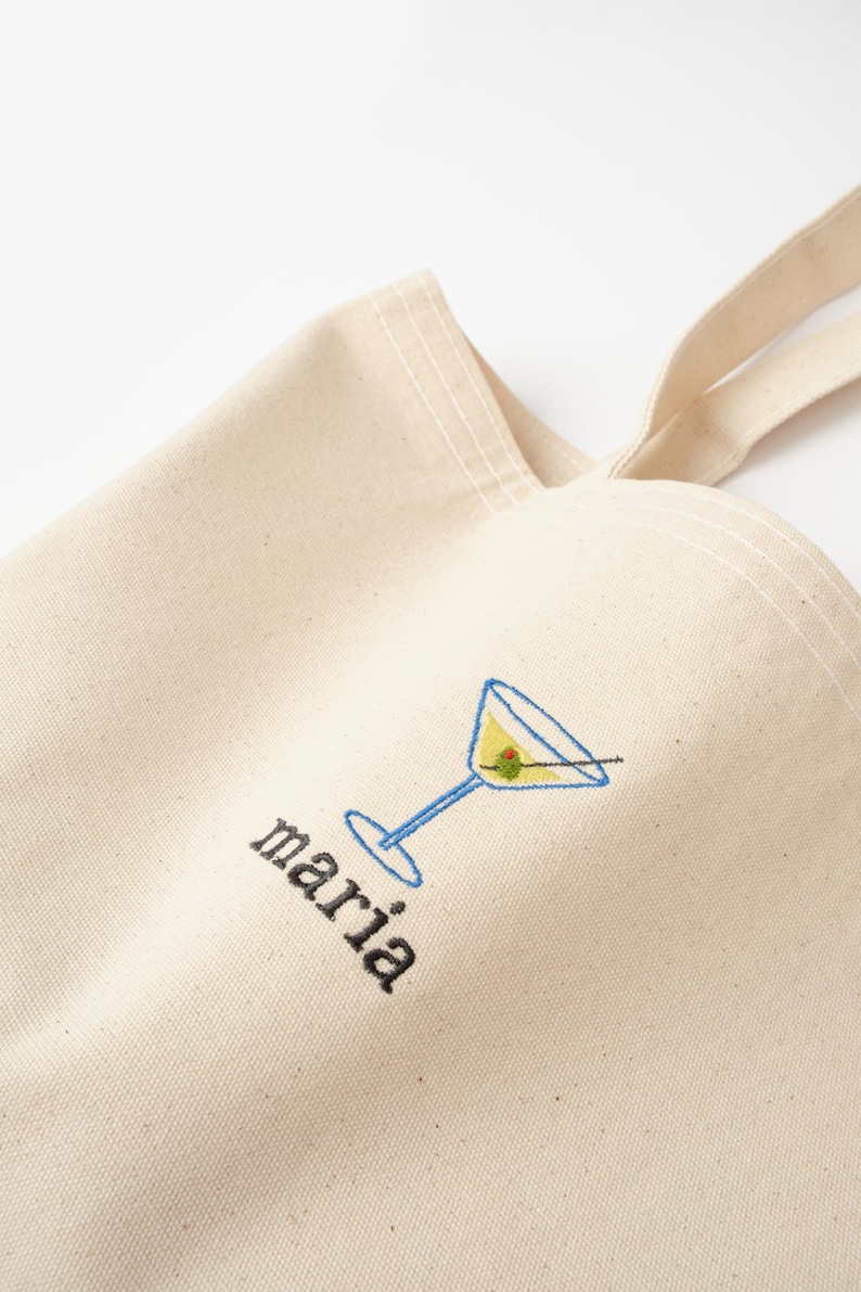 May include: A white canvas tote bag with a blue and yellow martini glass and olive embroidered on the front. The word "maria" is embroidered below the glass.