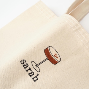 May include: A white canvas tote bag with a brown and black embroidered design of a cocktail glass. The bag has a black embroidered name "sarah" on the front.