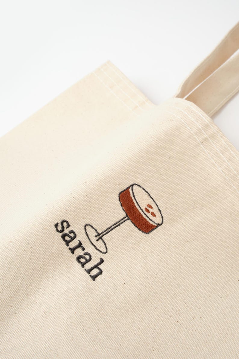 May include: A white canvas tote bag with a brown and black embroidered design of a cocktail glass. The bag has a black embroidered name "sarah" on the front.