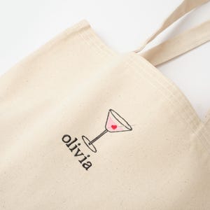 May include: A beige canvas tote bag with a black embroidered martini glass with a red heart inside. The bag has black embroidered text that says "olivia".
