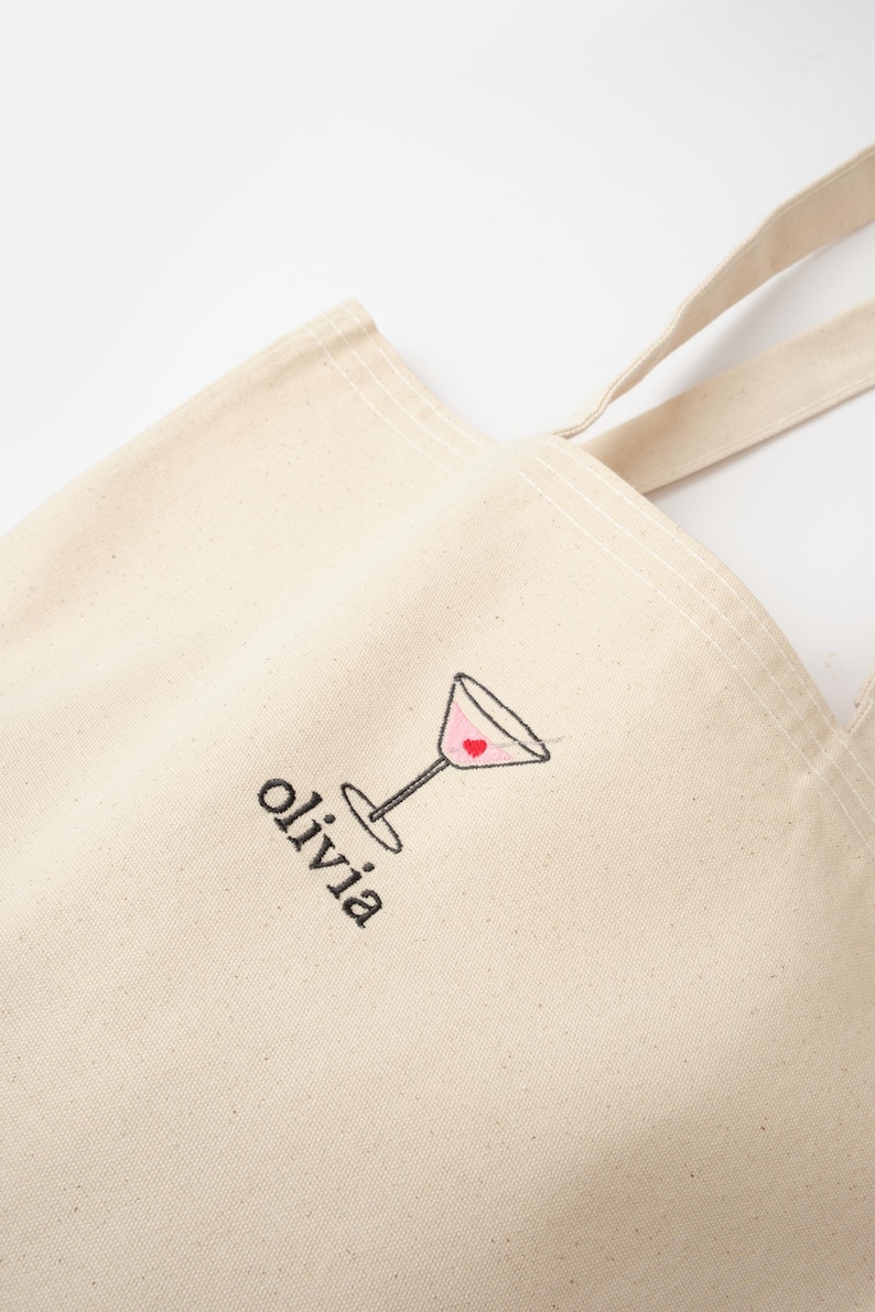 May include: A beige canvas tote bag with a black embroidered martini glass with a red heart inside. The bag has black embroidered text that says "olivia".