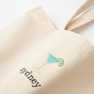 May include: A beige canvas tote bag with a blue and green embroidered cocktail and the word "sydney" in black embroidery.