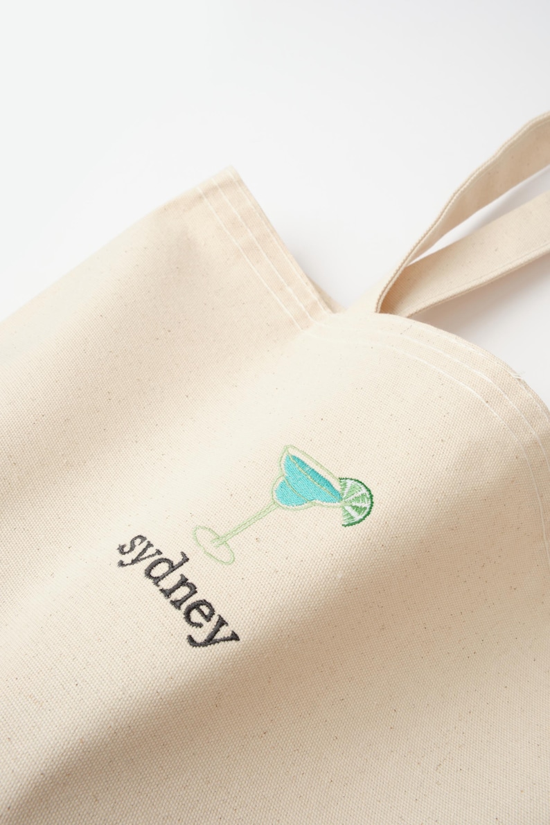 May include: A beige canvas tote bag with a blue and green embroidered cocktail and the word "sydney" in black embroidery.