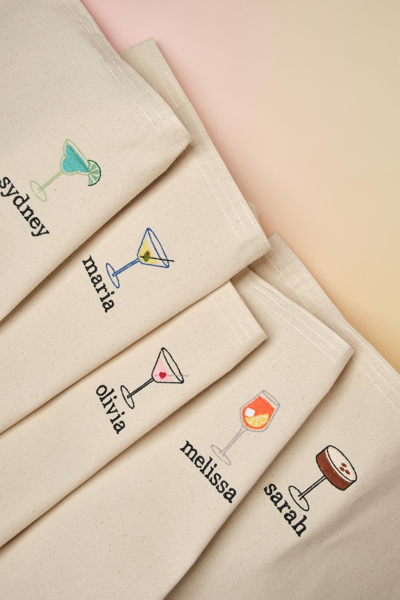 May include: Five white canvas tote bags with embroidered names and cocktail illustrations. The names are Sydney, Maria, Olivia, Melissa, and Sarah. Each bag features a different cocktail design.