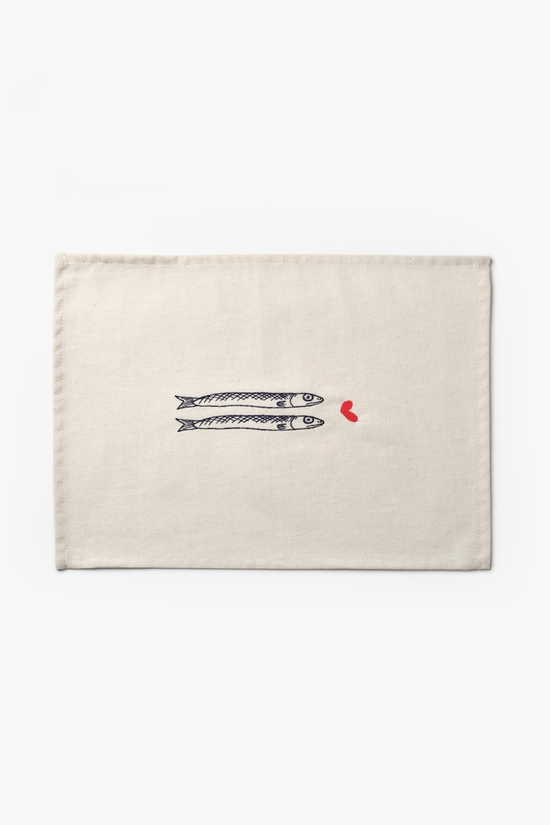 Puede incluir: Mantel individual color crema con un dise&ntilde;o caprichoso de dos peces estilizados y un coraz&oacute;n rojo. Los peces est&aacute;n dibujados en negro, contrastando con el fondo neutro. Una adici&oacute;n encantadora a cualquier mesa.