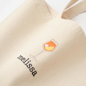 May include: A white canvas tote bag with a black embroidered design of a glass of orange juice and the word "melissa".