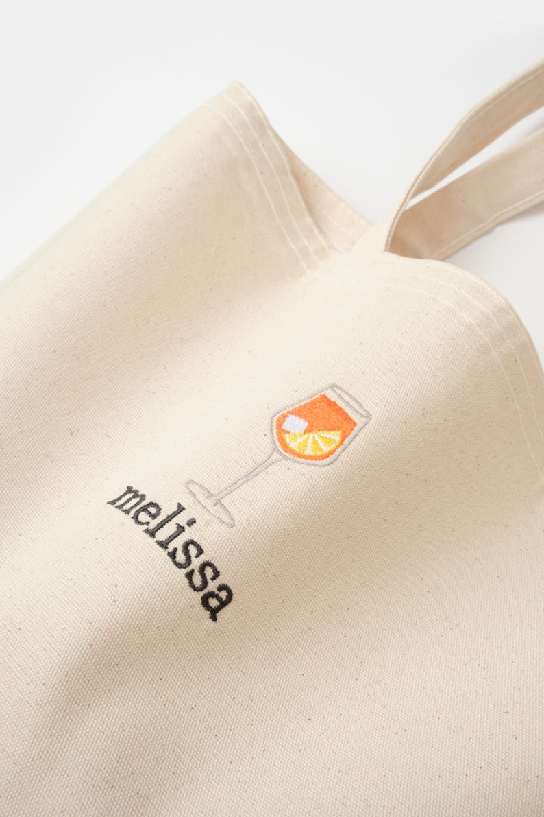 May include: A white canvas tote bag with a black embroidered design of a glass of orange juice and the word "melissa".