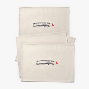 Puede incluir: Manteles individuales de color crema con un dise&ntilde;o caprichoso. Cada mantel individual presenta dos peces estilizados en tinta negra, con un peque&ntilde;o coraz&oacute;n rojo. Los manteles individuales son rectangulares y parecen estar hechos de tela natural.
