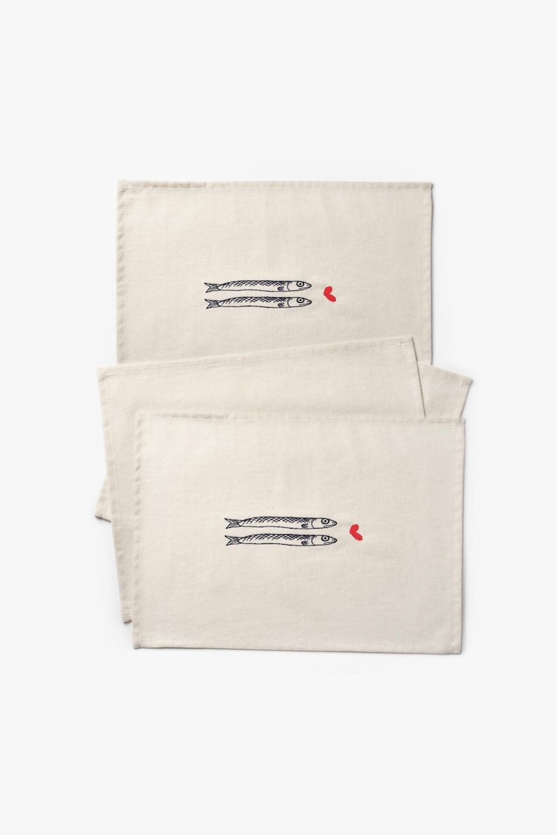 Puede incluir: Manteles individuales de color crema con un dise&ntilde;o caprichoso. Cada mantel individual presenta dos peces estilizados en tinta negra, con un peque&ntilde;o coraz&oacute;n rojo. Los manteles individuales son rectangulares y parecen estar hechos de tela natural.