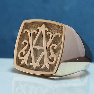 Könnte beinhalten: Ein goldener Siegelring mit einer quadratischen Fläche, auf der ein eingraviertes Monogramm der Buchstaben "A" und "S" zu sehen ist.