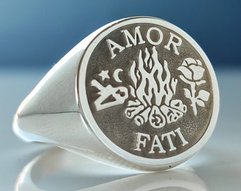 Könnte beinhalten: Ein silberner Siegelring mit kreisförmiger Oberfläche. Die Oberfläche zeigt die Worte "AMOR FATI" um ein zentrales Design aus Lagerfeuer, Rose, Mond und Stern. Der Ring ist hochglanzpoliert und reflektiert das Licht.