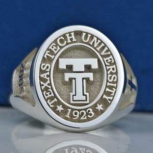 May include: A silver ring with an oval-shaped top featuring the words "Texas Tech University" and a stylized "T" in the center. The year "1923" is inscribed below the "T".