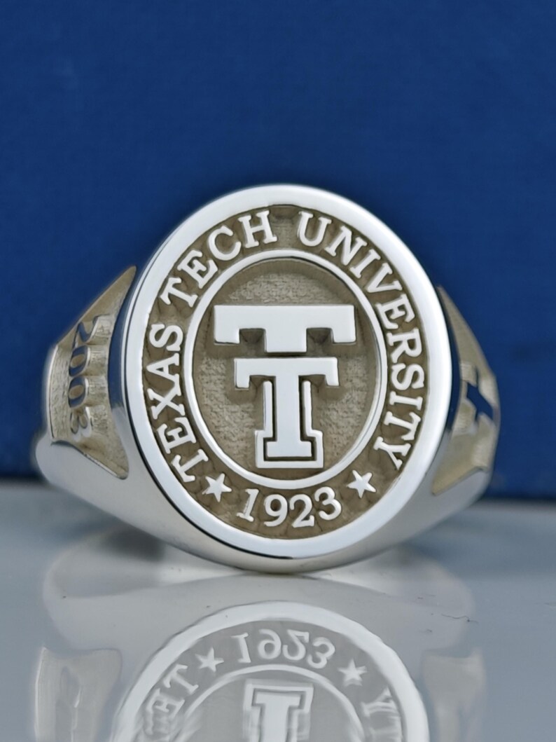 May include: A silver ring with an oval-shaped top featuring the words "Texas Tech University" and a stylized "T" in the center. The year "1923" is inscribed below the "T".