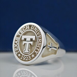 May include: A silver ring with a round, engraved top featuring the Texas Tech University logo and the year 1923. The ring has a cross design on the side.