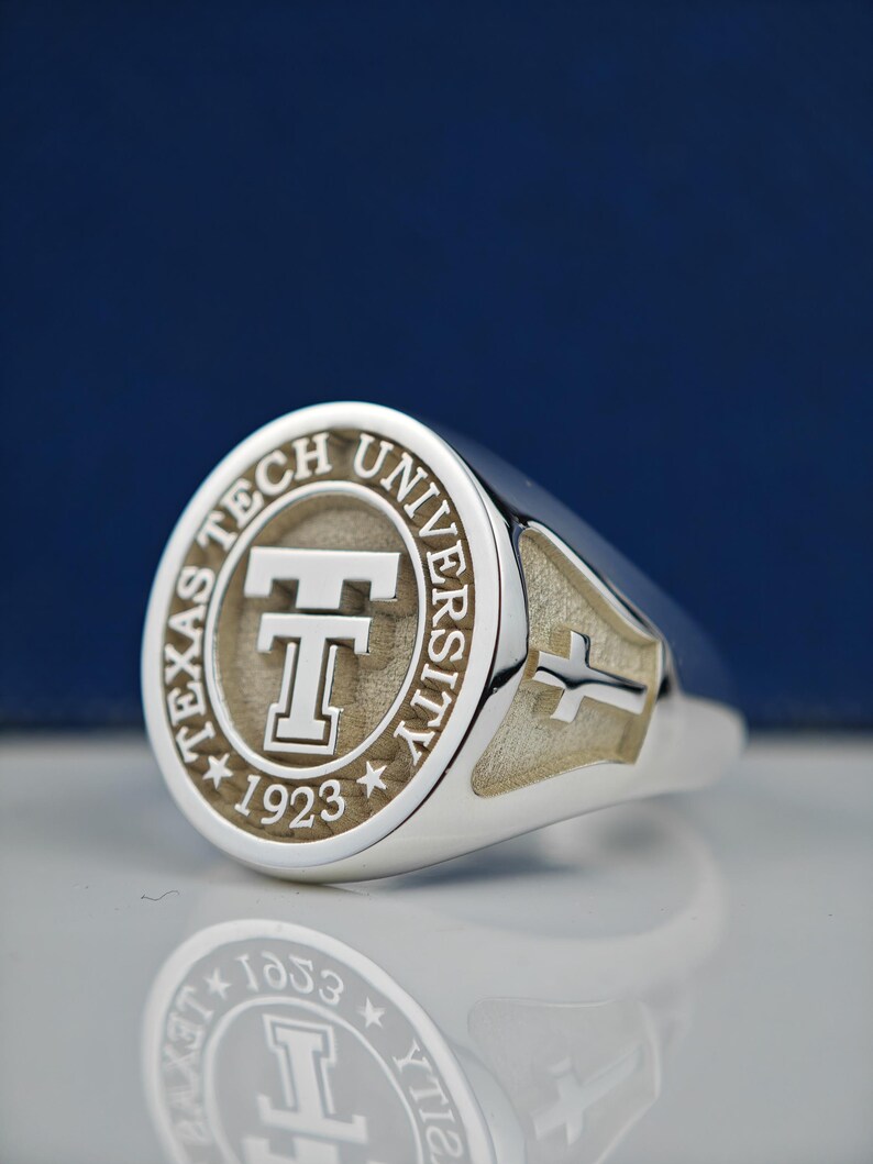 May include: A silver ring with a round, engraved top featuring the Texas Tech University logo and the year 1923. The ring has a cross design on the side.