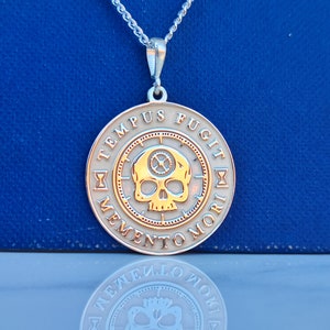 K&ouml;nnte beinhalten: Eine silberne Kettenkette mit einem runden Anh&auml;nger, der einen goldenen Totenkopf mit Zahnr&auml;dern und der Inschrift "TEMPUS FUGIT MEMENTO MORI" in einem Kreis zeigt.