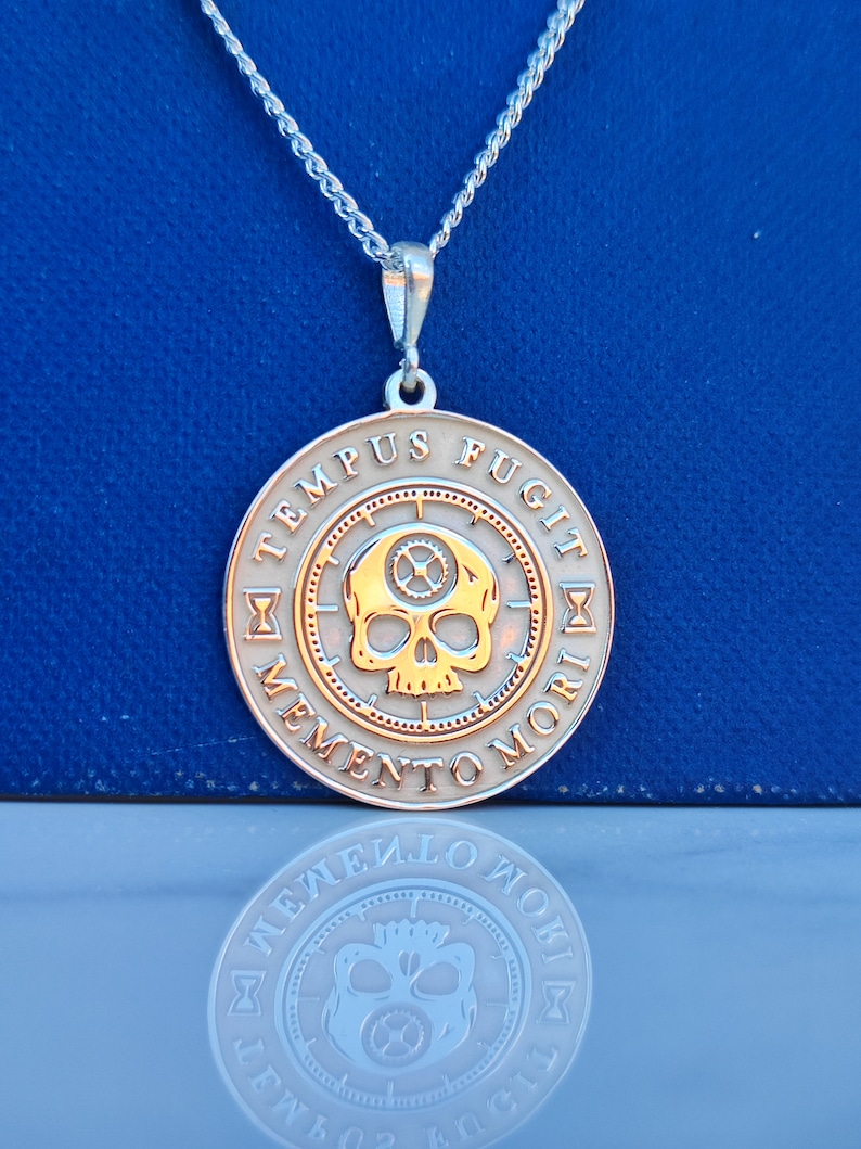 K&ouml;nnte beinhalten: Eine silberne Kettenkette mit einem runden Anh&auml;nger, der einen goldenen Totenkopf mit Zahnr&auml;dern und der Inschrift "TEMPUS FUGIT MEMENTO MORI" in einem Kreis zeigt.