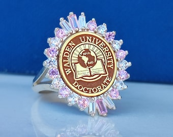 Könnte beinhalten: Ein silberner Ring mit einem braunen und goldenen runden Emblem, auf dem "Walden University" und "Doctorate" steht, mit einem pinkfarbenen und weißen Kristalldesign.