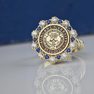 May include: A gold ring with a blue and white stone setting. The ring features the Auburn University seal with the text "Auburn University, Instruction, Research, Extension, Established 1856, 05.11.2025".