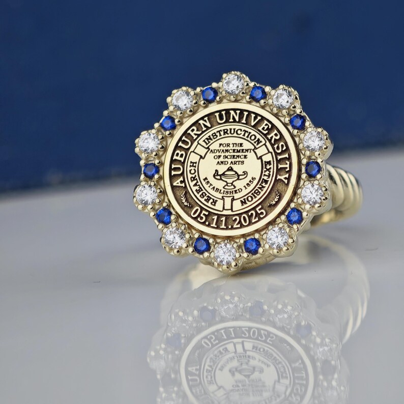 May include: A gold ring with a blue and white stone setting. The ring features the Auburn University seal with the text "Auburn University, Instruction, Research, Extension, Established 1856, 05.11.2025".