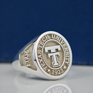 May include: A silver ring with a raised circular design featuring the words "Texas Tech University" and a large "T" in the center. The year "2003" is engraved on the side of the ring.