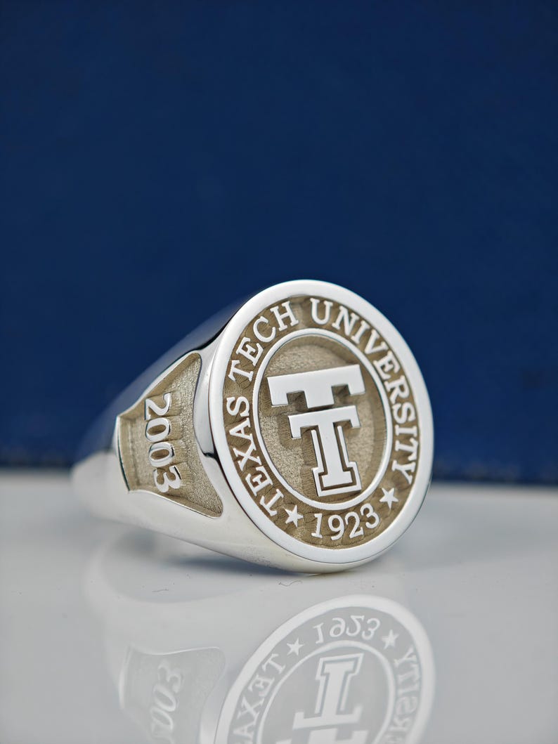 May include: A silver ring with a raised circular design featuring the words "Texas Tech University" and a large "T" in the center. The year "2003" is engraved on the side of the ring.
