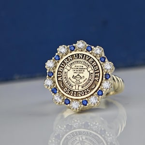 May include: A gold ring with a blue and white stone setting. The ring features the Auburn University seal with the text "Auburn University, Instruction, Research, Extension, For the Advancement of Science and Arts, Established 1856, 05.11.2025".