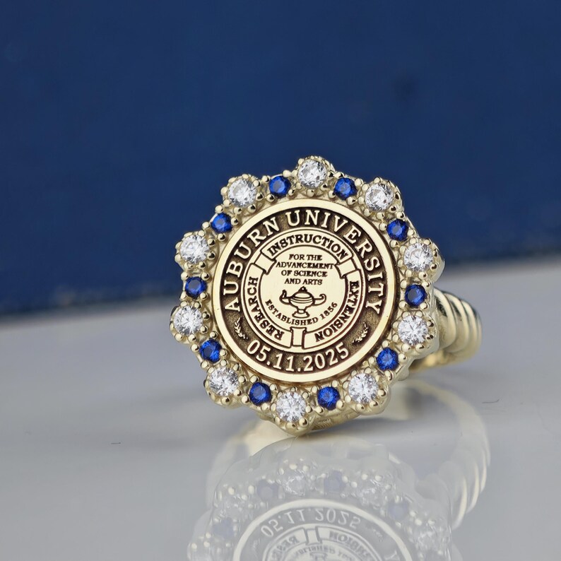May include: A gold ring with a blue and white stone setting. The ring features the Auburn University seal with the text "Auburn University, Instruction, Research, Extension, For the Advancement of Science and Arts, Established 1856, 05.11.2025".
