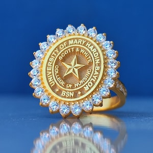 May include: Gold ring with a circular design featuring the inscription "University of Mary Hardin-Baylor Scott & White College of Nursing BSN". The ring is adorned with a star and surrounded by small, clear gemstones.