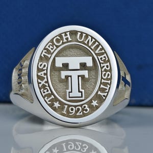 May include: A silver ring with a raised oval design featuring the words "Texas Tech University" and a large "T" in the center. The year "1923" is inscribed below the "T" and two stars are on either side of the year. The year "2008" is inscribed on the side of the ring.