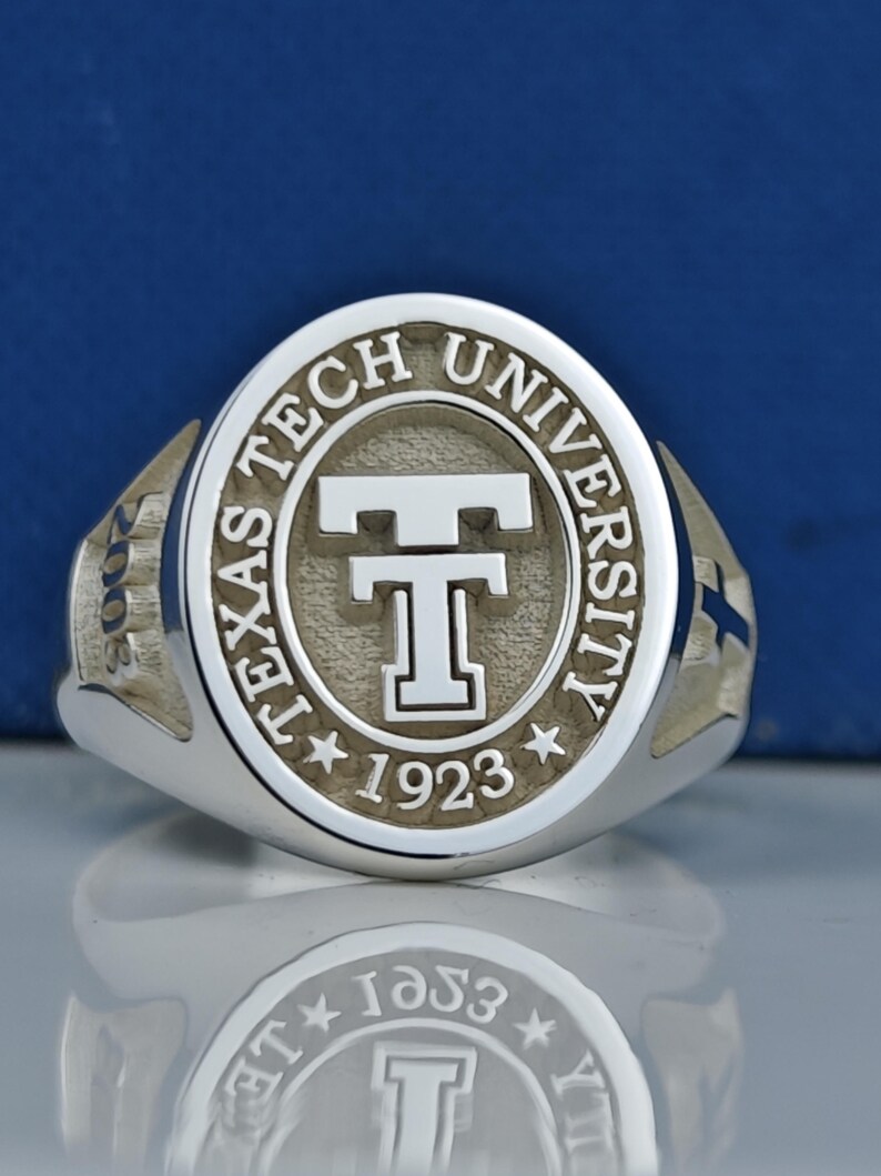 May include: A silver ring with a raised oval design featuring the words "Texas Tech University" and a large "T" in the center. The year "1923" is inscribed below the "T" and two stars are on either side of the year. The year "2008" is inscribed on the side of the ring.