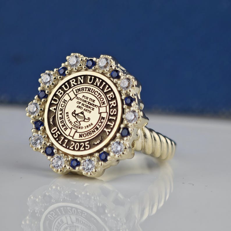 May include: A gold ring with a blue and white stone setting. The ring features the Auburn University seal with the words "Auburn University" and "Instruction, Research, Extension" around the edge. The seal also includes the words "For the Advancement of Science and Arts" and "Established 1856".