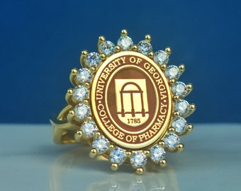 Könnte beinhalten: Ein goldener Ring mit einem braunen, ovalen Emblem, das das Logo des College of Pharmacy der University of Georgia und das Jahr 1785 zeigt. Das Emblem ist von einem Kreis aus klaren, funkelnden Steinen umgeben.