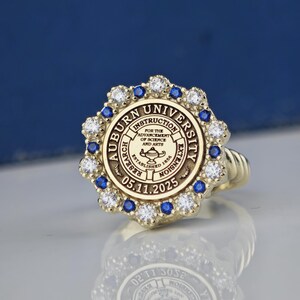 May include: A gold ring with a blue and white stone setting. The ring features the Auburn University seal with the text "Auburn University" and "05.11.2025".