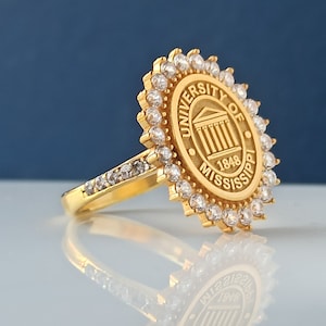 May include: A gold ring with a circular design featuring the University of Mississippi logo and the words "University of Mississippi 1848" surrounded by clear stones.