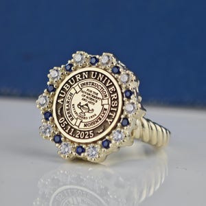 May include: A gold ring with a blue and white stone setting. The ring features the Auburn University seal with the text "Auburn University, Instruction, Research, Extension, Established 1856, 05.11.2025".