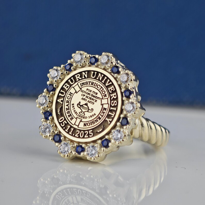 May include: A gold ring with a blue and white stone setting. The ring features the Auburn University seal with the text "Auburn University, Instruction, Research, Extension, Established 1856, 05.11.2025".