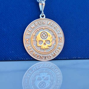 K&ouml;nnte beinhalten: Eine silberne Halskette mit einem runden Anh&auml;nger, der einen goldenen Sch&auml;del mit Zahnr&auml;dern im Inneren zeigt. Der Anh&auml;nger tr&auml;gt die lateinische Inschrift "Tempus Fugit Memento Mori" am Rand.