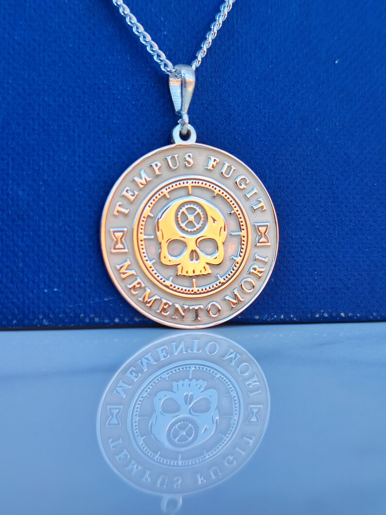K&ouml;nnte beinhalten: Eine silberne Halskette mit einem runden Anh&auml;nger, der einen goldenen Sch&auml;del mit Zahnr&auml;dern im Inneren zeigt. Der Anh&auml;nger tr&auml;gt die lateinische Inschrift "Tempus Fugit Memento Mori" am Rand.